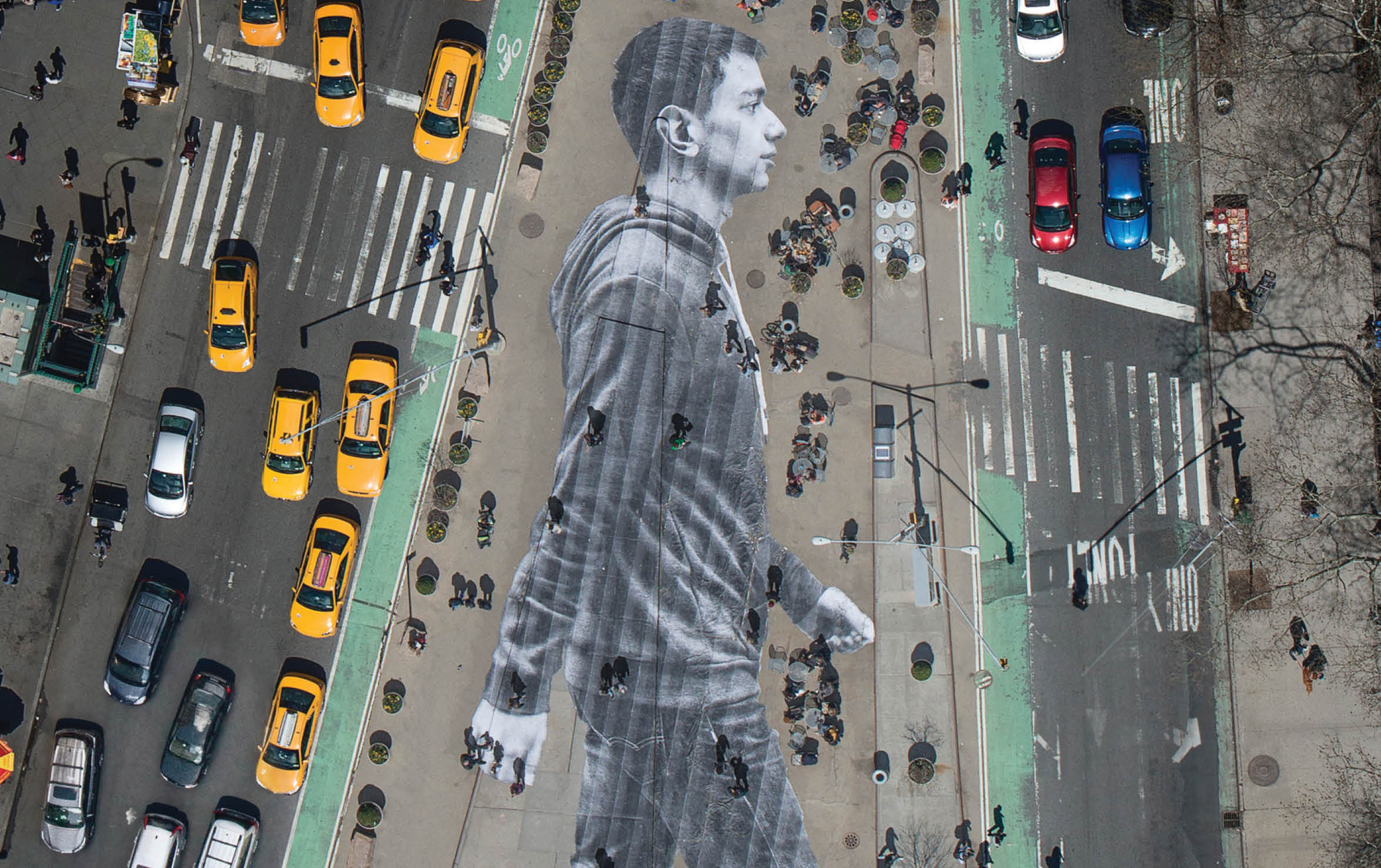 What you see on the cover of this week s New York Times Magazine is real  The image is by the French artist JR, who is known for pasting giant photographs on urban surfaces all over the world  About six weeks ago, we met with JR to discuss a project for our Walking New York issue  Lately, JR s work has been preoccupied with the theme of immigration  An installation at Ellis Island features large archival photographs of immigrants displayed throughout the island s abandoned hospital   For this project, we decided we d photograph recent immigrants and paste their images on the city s streets, where they and other immigrants are often invisible  The pasting would be made in the heart of Manhattan, on Flatiron plaza, the triangle of pavement between Fifth Avenue, Broadway and East 23rd Street   Earlier this month, JR photographed 16 people who arrived in New York within the last year and made their portraits while they walked down the streets of NoLIta  (Some of these portraits can been seen here ) For the cover, we zeroed in on Elmar Aliyev, a 20-year-old waiter at Old Baku, an Azerbaijani restaurant on Ditmas Avenue in Brooklyn  Elmar immigrated to the United States from Azerbaijan last August after he won the green-card lottery  He likes to walk in Central Park and around the Plaza, locations from his favorite movie,  Home Alone 2   JR liked the way Aliyev swung his arms when he walked   Aliyev s picture was then printed on 62 strips of paper  On April 11, JR and his 20-person crew took these strips to Flatiron plaza and began affixing them to the ground at 4 a m  The pasting took about three and a half hours and resulted in a 150-foot-tall image of Aliyev striding eastward  The sun came up  Pedestrians began to wander over Aliyev  Just as JR had predicted, they often walked right over him without even noticing   To make out the image, you had to be high above  In the middle of the day, when the angles of shadow were favorable, JR went up in a helicopter and photographed the pasting, with all the cheerful and untidy street life of an early spring day in New York City happening on and around it   That image is the one you see on the cover of the magazine — a gloriously massive portrait of one of the city s 3 1 million immigrants, unseen by many of the people passing him on the street 